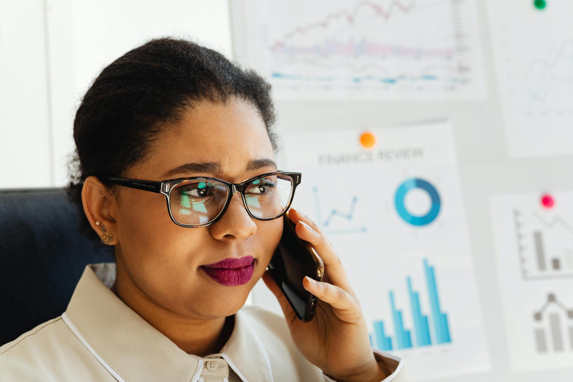Focused woman in glasses discusses financial graphs on phone inside office.